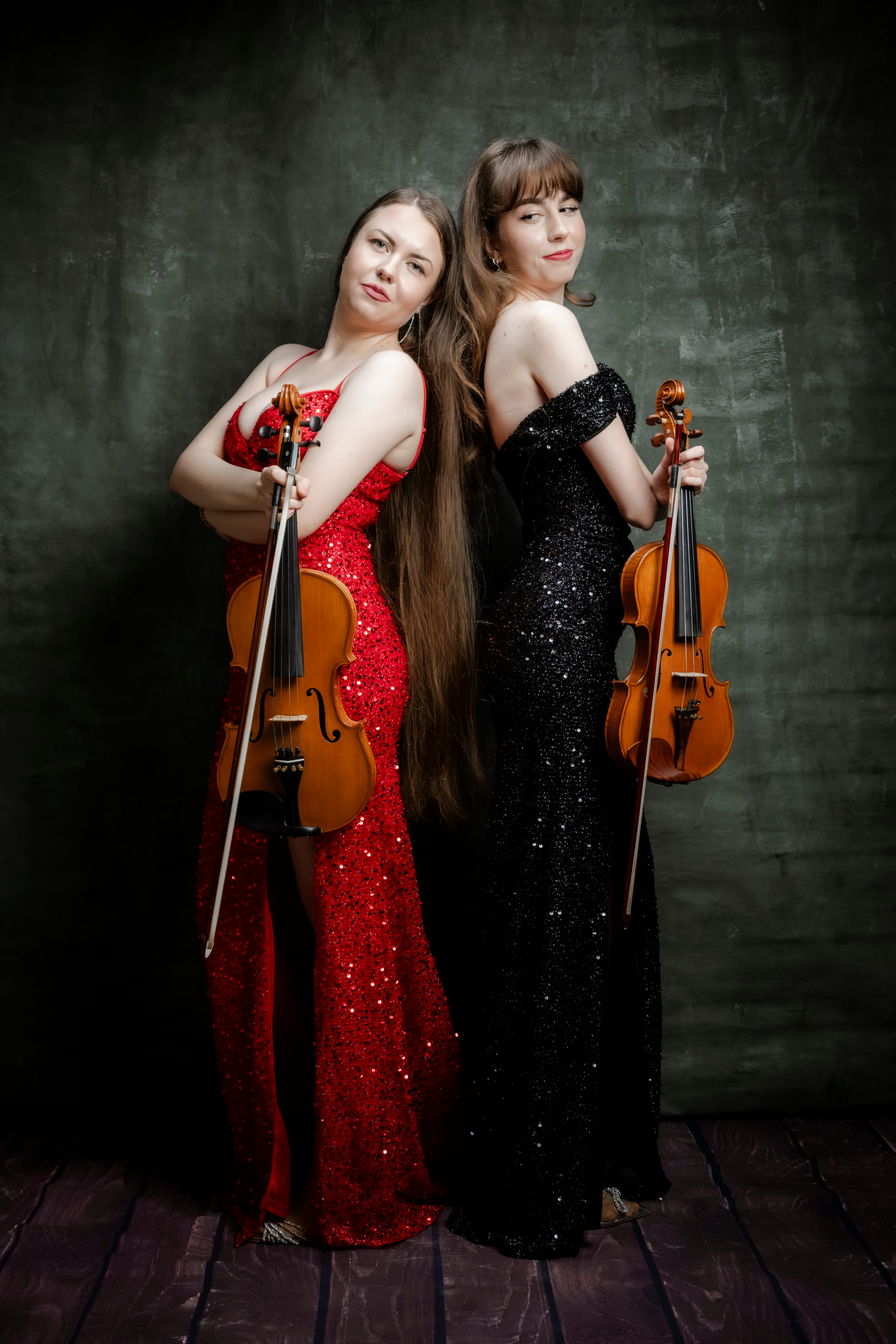 Duo-ul Sistrings stând spate în spate, în rochii roșie și neagră, cu vioară și violă